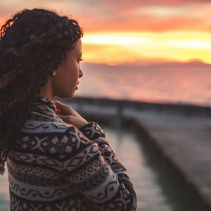 Thoughtful evening mood with a young Afro American woman standing on the promenade by the lake and looking towards the water and the setting sun. Burning sky